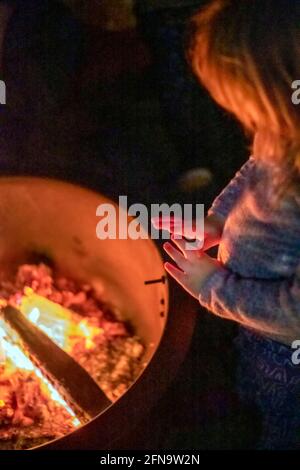 Il bambino riscalda le mani di fronte ad una fossa di fuoco Foto Stock