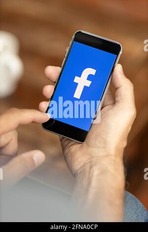 Uomo che tiene uno smartphone in un bar. Logo Facebook sul display. Messa a fuoco selettiva. Foto Stock