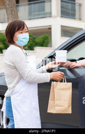 Donna asiatica che consegna cibo da asporto a un cliente in un'auto: Concentrazione selettiva. Concetto aziendale e di sicurezza. Foto Stock