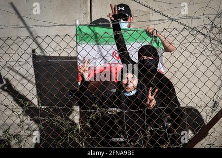 Aadyisseh, Libano. 15 maggio 2021. I sostenitori del movimento hezbollah pro-Iran libanese, detengono una bandiera iraniana e lampeggiano i segni della vittoria mentre si trovano vicino a un muro eretto dall'esercito israeliano lungo il confine libanese-israeliano, durante una protesta nel villaggio meridionale di Aadyisseh in solidarietà con i palestinesi, in mezzo all'escalation della violenza israelo-palestinese. Credit: Marwan Naamani/dpa/Alamy Live News Foto Stock