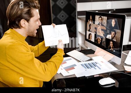 Elegante uomo d'affari freelance in camicia gialla seduto alla scrivania e al PC, che tiene una conferenza online tramite l'app. Manager che mostra i grafici finanziari del team Foto Stock
