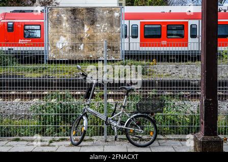 Una bicicletta parcheggiata in una stazione ferroviaria con vista di un cartellone inutilizzato, dei binari e di una fermata del treno rosso, Foto Stock