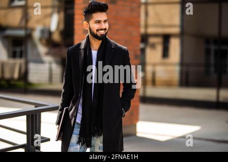 Giovane indiano che indossa abiti demi-season per strada. Giovane ragazzo con bearded e moderno acconciatura con cappotto, jeans blu e t-shirt contro Urban bl Foto Stock