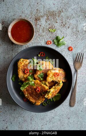 Frittelle di mais indonesiane fatte in casa (Perkedel Jagung o Bakwan Jagung) servite con salsa di peperoncino dolce su piatto nero e sfondo blu testurizzato, copia Foto Stock