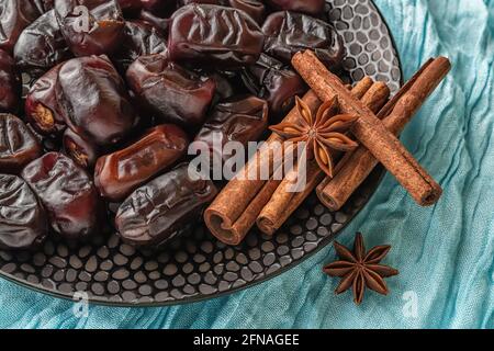 Deliziosi datteri iraniani secchi, pochi bastoncini di cannella e anice stellato su piastra di ceramica nera su tela turchese. Cibo dolce vegetariano sano. Foto Stock