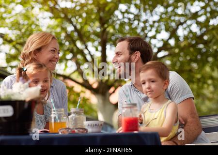 felici genitori caucasici che si guardano, sorridenti, con i loro figli in giri, seduti all'aperto Foto Stock