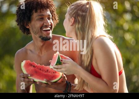 giovane coppia multietnica che si guarda l'un l'altro, sorridendo, ridendo, mentre si mangia il cocomero Foto Stock