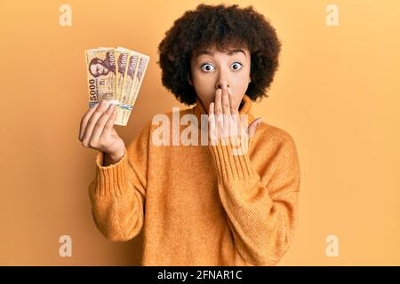 Giovane ragazza ispanica che tiene 5000 banconote forinte ungheresi che coprono la bocca con la mano, scioccata e impaurita per errore. Sorpresa espressione Foto Stock