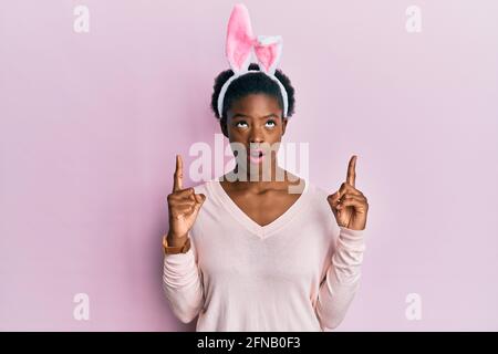 Giovane afroamericana ragazza che indossa le orecchie carine coniglietto di pasqua stupito e sorpreso guardando in su e puntando con le dita e le braccia sollevate. Foto Stock