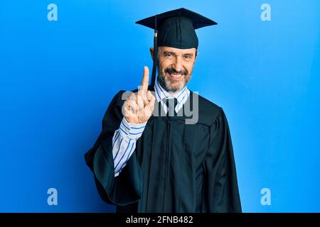 Uomo ispanico di mezza età che indossa un cappuccio di graduazione e un abito da cerimonia mostrando e puntando verso l'alto con il dito numero uno mentre sorride sicuro e felice. Foto Stock