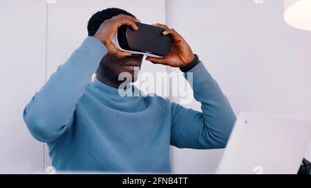 Uomo nero che indossa un bicchiere di realtà virtuale seduto alla scrivania con il suo laptop. Foto di alta qualità Foto Stock