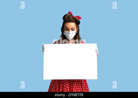 Combatte il coronavirus. Giovane signora pinup in maschera di faccia che tiene banner vuoto con mockup per il tuo disegno su sfondo blu Foto Stock