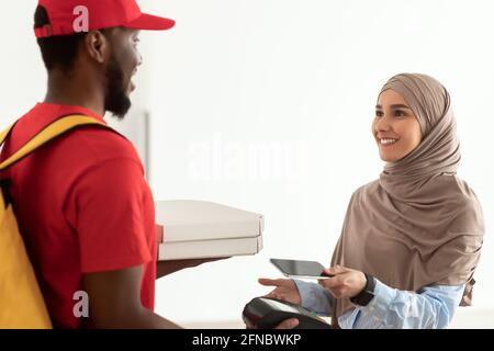 Addetto alla consegna nero che tiene la scatola per pizza e il terminale pos Foto Stock