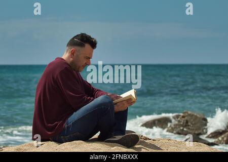 uomo seduto sulla sabbia che legge un libro sul mare con il mare sullo sfondo Foto Stock