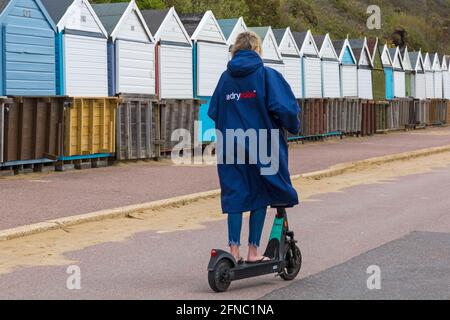 Bournemouth, Dorset, Regno Unito. 16 maggio 2021. Meteo del Regno Unito: Clima misto con docce a pioggia e un po' di sole sulle spiagge di Bournemouth, mentre la gente si dirige verso il mare per l'aria fresca e l'esercizio fisico. Donna che guida Beryl scooter Beryl e-scooter escooter e scooter scooter elettrico lungo la passeggiata, indossa abiti asciutti accappatoi asciutti dryrobe - vista posteriore. Crediti: Carolyn Jenkins/Alamy Live News Foto Stock