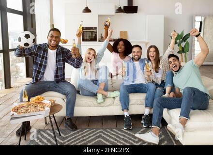 Amici multietnici entusiasti che bevono birra e mangiano spuntini, guardando la partita di calcio in TV a casa Foto Stock