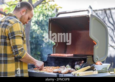 Uomo all'aperto con barbecue. Giovane uomo che grigliano spiedini di carne, bistecca di carne, melanzane e funghi su una griglia a gas. L'attenzione selettiva è rivolta alla salsiccia e alla bistecca. Foto Stock
