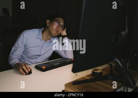 Giovane uomo d'affari asiatico che lavora overworking in ufficio corporativo. Lavoratore maschile di ufficio che pensa e si sta stress durante le ore di straordinario di notte Foto Stock