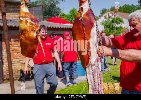 Aradac, Vojvodina, Serbia - 21 maggio 2016: Due prosciutti di maiale crudi sono appesi sopra nel cortile del villaggio, macellaio sta tagliando la carne. Foto Stock