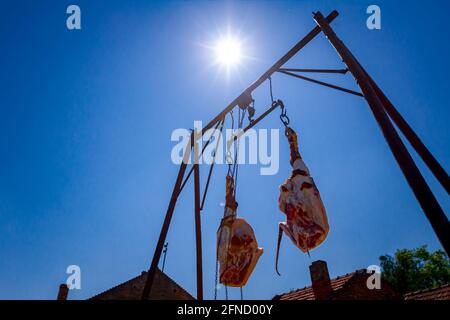 Sparato dal basso su due prosciutti di maiale freschi e crudi agganciati a ganci, appesi su un supporto metallico. Vista sui raggi del sole e il cielo blu. Foto Stock