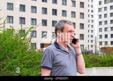Uomo caucasico adulto anziano in una T-shirt che parla su uno smartphone in città in primavera o in estate sullo sfondo degli edifici. Messa a fuoco selettiva. Foto Stock