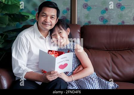 Le piccole figlie asiatiche si congratulano con papà e gli danno una cartolina E confezione regalo con sorridente e hugging.Concept di felice padre giorno Foto Stock