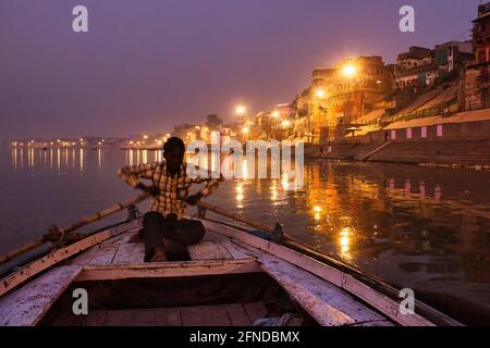 Varanasi, India - 01 novembre 2016: Un uomo che remi una pagaia di legno su una barca prima dell'alba nel fiume ganges contro loca illuminata città Santa varanasi Foto Stock