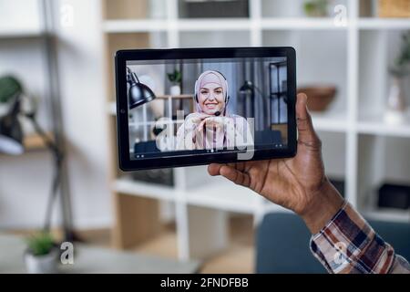 Primo piano della mano maschile che tiene la tavoletta digitale con la donna musulmana in hijab sullo schermo. Giovane uomo africano che parla con la ragazza attraverso la videochiamata durante la pandemia. Foto Stock