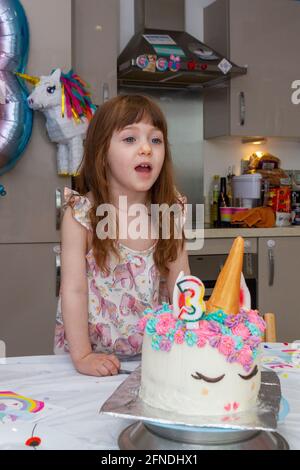 Una ragazza carina, dai capelli marroni in una cucina che soffia candele di una torta di compleanno decorata come unicorno con una candela numero tre con un pinhata in th Foto Stock