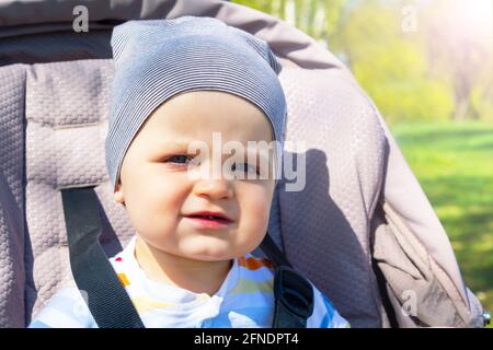 Adorabile bambino caucasico felice seduto in passeggino grigio nel parco. Giorno d'estate. Il bambino viene stralciato in passeggino durante la passeggiata. Foto Stock