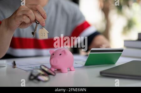 Risparmio di denaro per la prima casa o casa ipotecario concetto di prestito, uomo tenendo in legno casa catena chiave con bianco piggy banca su tavola di legno con spazio copia Foto Stock