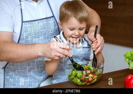 Il figlio e il padre dai capelli biondi preparano insieme in cucina un'insalata di verdure fresche e colorate. Entrambi indossavano grembiuli blu. Foto Stock