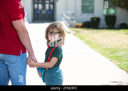 Ritratto di allievo felice nerd che tiene gli insegnanti a mano. Scolaro che va a scuola con il padre. Genitore con bambino di fronte alle porte della scuola. Foto Stock