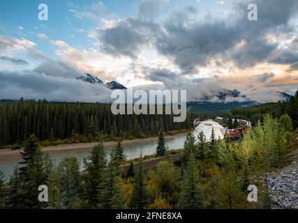 Cloudy Rocky Mountains al tramonto, Morant's Curve, treno in lontananza nella curva, rive boscose a Bow River, Bow Valley, Banff National Park, Alberta Foto Stock