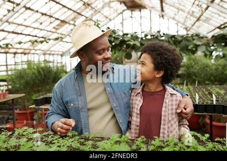 Uomo africano abbracciando il bambino e parlandogli durante la piantagione piantine in serra Foto Stock