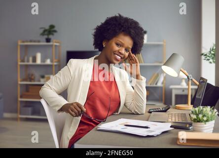 Felice rilassata e sicura giovane donna d'affari nera seduta alla scrivania dell'ufficio Foto Stock