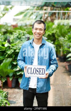 Giovane uomo allegro che tiene il segno chiuso quando in piedi nel giardinaggio centro Foto Stock