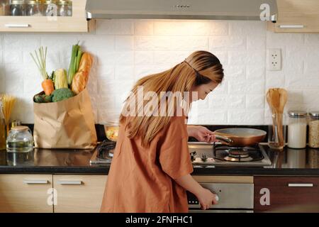 Giovane donna che si accende la stufa a gas per friggere le verdure o preparare la salsa di pasta per la cena, vista dal retro Foto Stock