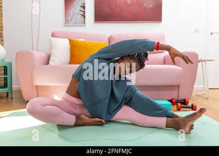 Donna nera seduta sul tappetino e gambe che si allungano mentre si fa curva laterale durante l'allenamento a casa Foto Stock