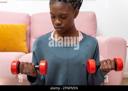 Donna sportiva nera concentrata che si esercita con manubri sul tappetino durante l'allenamento a casa Foto Stock