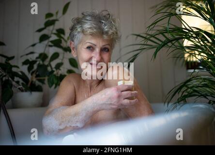 Donna anziana contented che si lava nella vasca da bagno a bolle a casa, guardando la macchina fotografica. Foto Stock