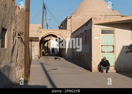 Uomo anziano che si zola nel tardo pomeriggio sole mentre un gatto attraversa una strada nel villaggio Mohammadieh vicino a Nain, Contea di Nain, Provincia di Isfahan, Iran Foto Stock