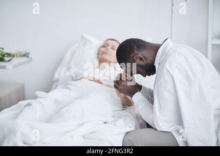 Uomo triste africano seduto di fronte al letto d'ospedale con sua moglie e preoccupato per la sua salute Foto Stock