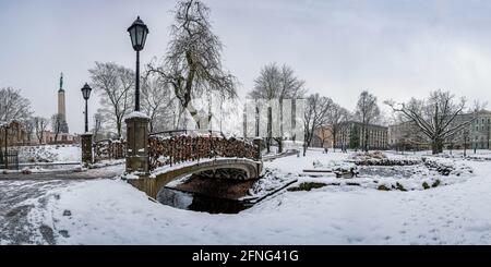 Vista del parco Bastion Hill e del Monumento alla libertà a riga, Lettonia. Paesaggio invernale in un parco nevoso con un bellissimo piccolo ponte con serrature sullo stagno. Foto Stock