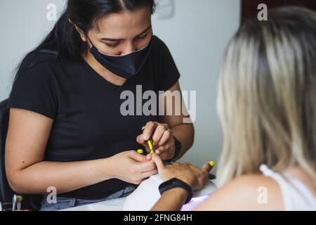 Crop femmina etnia non riconoscibile master in maschera sterile applicando giallo polacco su chiodi di cliente anonimo in salone di bellezza Foto Stock