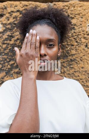 Donna afroamericana senza emozioni in camicia bianca che copre metà faccia con la mano e osservi la macchina fotografica contro il ruvido irregolare parete Foto Stock