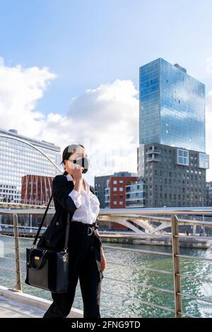 Imprenditore femminile di etnia in abbigliamento formale e maschera facciale in tessuto con la mano in tasca che parla sul cellulare mentre si guarda via in città Foto Stock