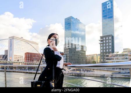 Imprenditore femminile di etnia in abbigliamento formale e maschera facciale in tessuto con la mano in tasca che parla sul cellulare mentre si guarda via in città Foto Stock