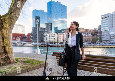 Sorridente imprenditore etnico femminile appoggiato su una panca di legno che parla sopra cellulare mentre si guarda via sulla panchina della città contro lo scooter Foto Stock