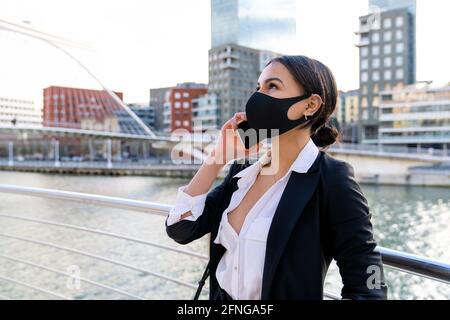 Imprenditore femminile di etnia in abbigliamento formale e maschera facciale in tessuto con la mano in tasca che parla sul cellulare mentre si guarda via in città Foto Stock